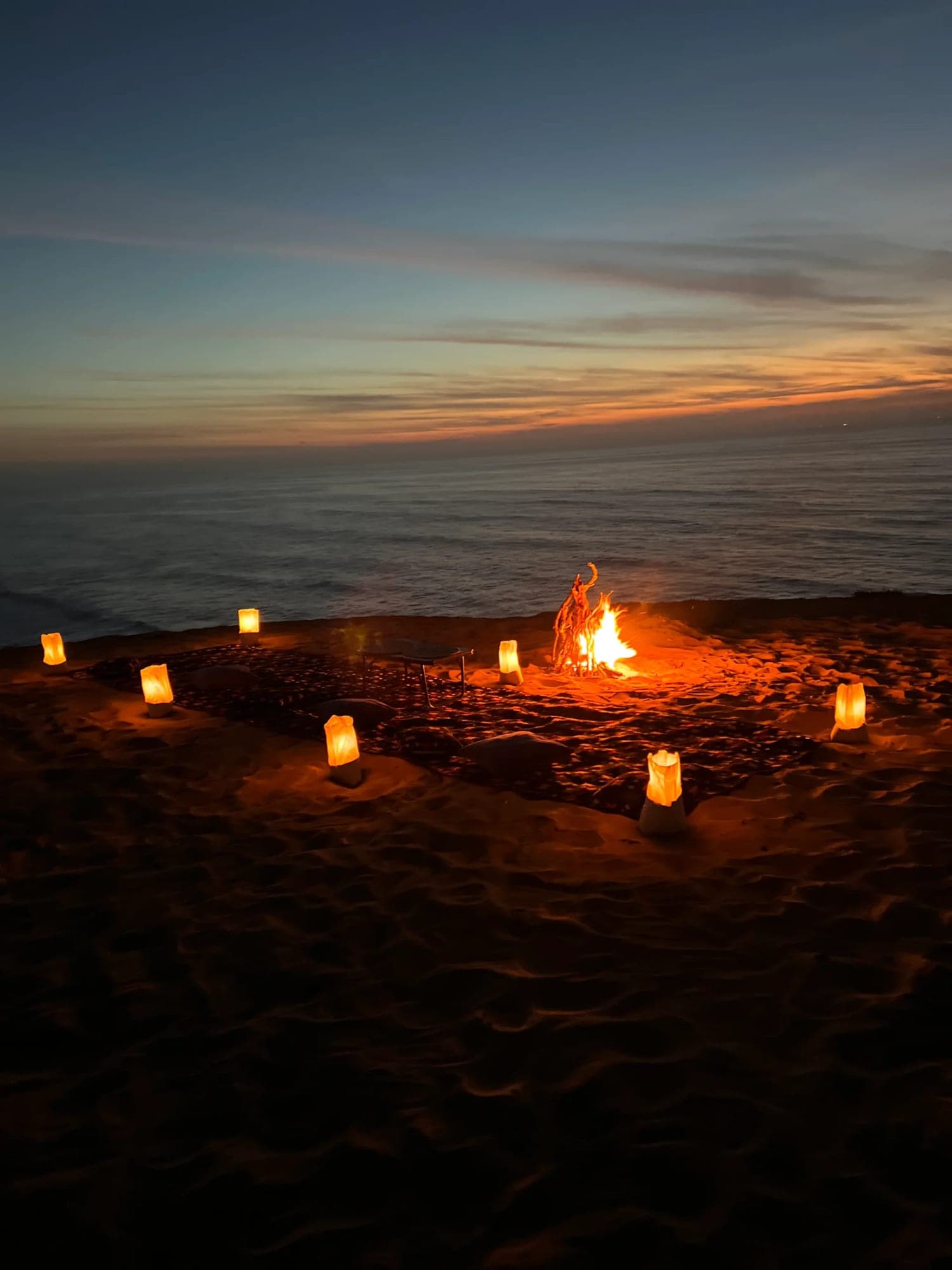 Dinner setting with rugs, lanterns and a campfire on the sand above the ocean at dusk near Timlalin