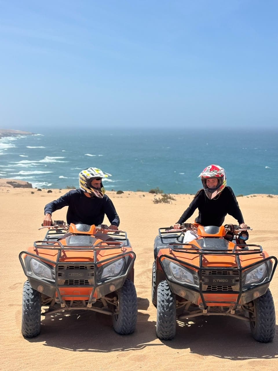 Two smiling riders in helmets on orange quad bikes on a Timlalin dune above the Atlantic Ocean