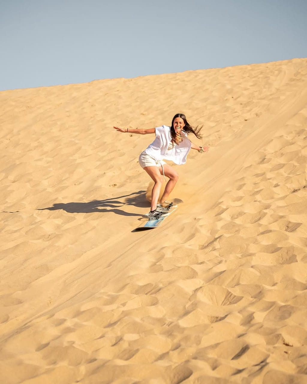 Smiling visitor sandboarding down a steep golden dune face at Timlalin