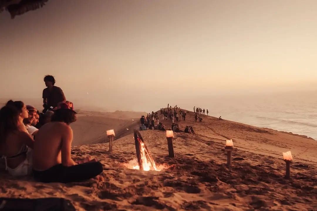 Visitors gathered around a campfire and candles on a dune ridge above the ocean at sunset near Timlalin