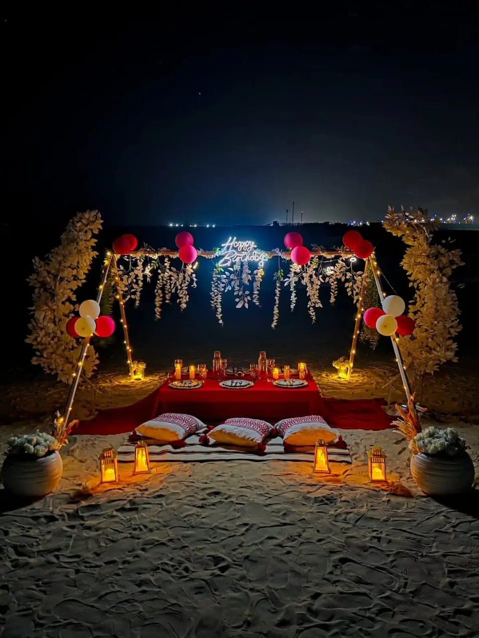Birthday celebration setup on the sand with a low table, cushions, lanterns and balloons under a lit arch at night